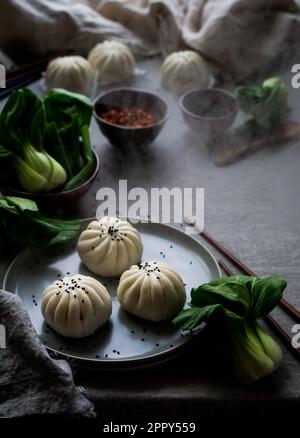 Panini al vapore cinesi guarniti con sesamo nero su un piatto con bastoncini, bok choi e salse sullo sfondo. Il vapore sta emanando dai panini. Foto Stock