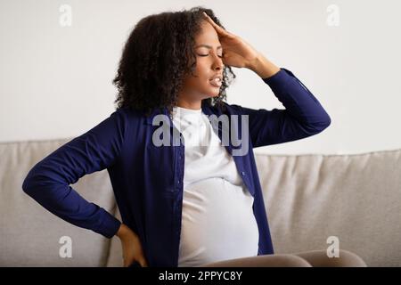 Triste giovane afroamericana donna incinta con pancia in casual sedersi sul divano, che soffre di emicrania Foto Stock