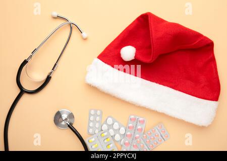 Medicina natale nuovo anno di concetto durante il coronavirus covid tempo. Pillole mediche con cappello di santa e stetoscopio su sfondo beige. Vista dall'alto. Foto Stock