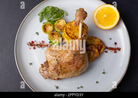 Confit di coscia d'anatra con germogli di Bruxelles, patate al forno, timo e arancia. Cucina francese tradizionale. Messa a fuoco selettiva, primo piano, sfondo nero. Foto Stock