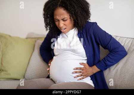 Giovane donna incinta afroamericana disperata siede sul divano che soffre di contrazioni, urlando Foto Stock