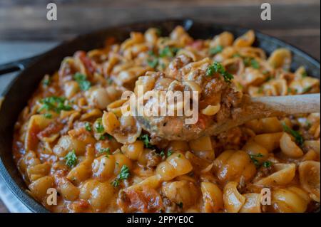 Macinare il manzo con gusci di panna, formaggio e pasta in una padella di ghisa con cucchiaio di legno Foto Stock