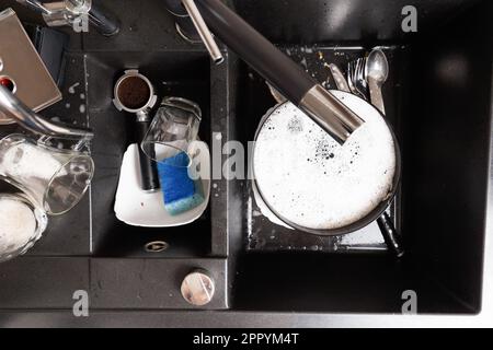 Concetto di lavaggio delle stoviglie. Piatti sporchi con schiuma di detergente in un lavello in pietra nera nella vista dall'alto della cucina. Bicchieri, tazze, piatti, posate sporchi Foto Stock