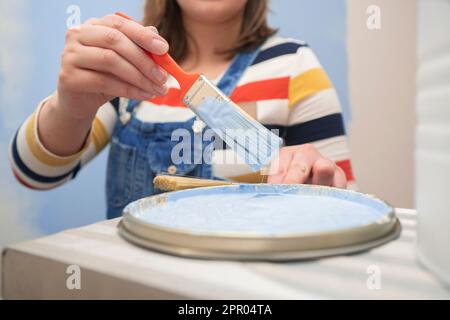 Primo piano della mano della donna indossando tute, togliendo una spazzola con vernice blu da un vassoio con vernice con sfondo a parete semi-dipinto Foto Stock