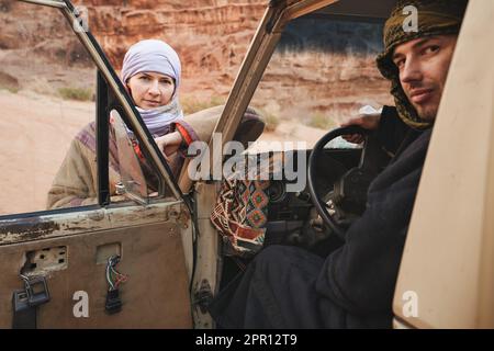 Giovane donna in camice beduino tradizionale - bisht - e foulard, seduto accanto al vecchio veicolo 4WD, guardando sopra la porta aperta, sfocato giovane uomo anche wea Foto Stock