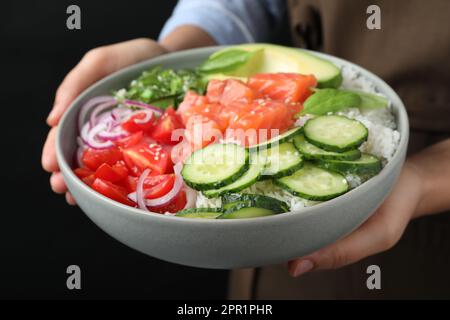 Donna che tiene deliziosa ciotola poke con salmone e verdure su sfondo nero, primo piano Foto Stock