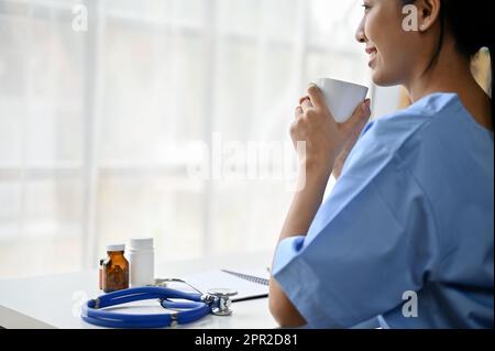 Vista laterale di un giovane studente di medicina asiatica seduto nella sala studio e con una tazza di caffè. Foto Stock