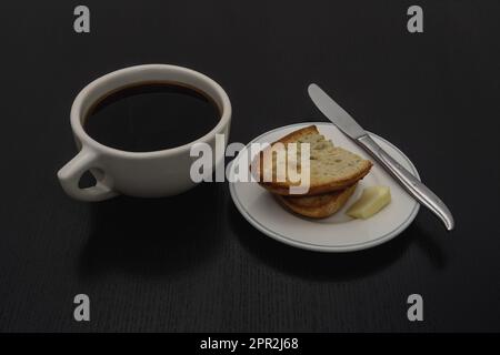 Una classica tazza di caffè nero in stile americano bianco si trova accanto a un piccolo piatto di pane tostato, burro e un coltello. Foto Stock