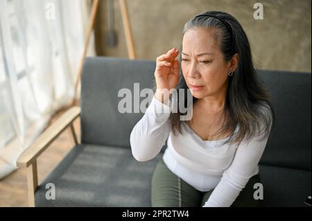 Donna anziana asiatica stressata che soffre di mal di testa o emicrania mentre pensiva qualcosa mentre si siede sul divano a casa. Foto Stock