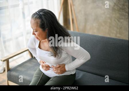 Malessere donna anziana asiatica affetta da mal di stomaco, intossicazione alimentare, gastrite o dolore addominale, sensazione di disagio, tenendo la pancia mentre si siede Foto Stock