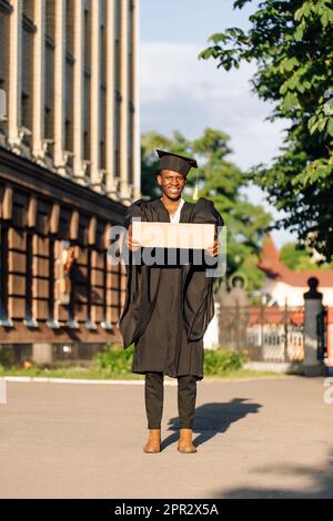 Giovane uomo nero disoccupato in piedi con poster di cartone vuoto per strada in cerca di lavoro. Studente universitario o universitario laureato in abito di laurea Foto Stock