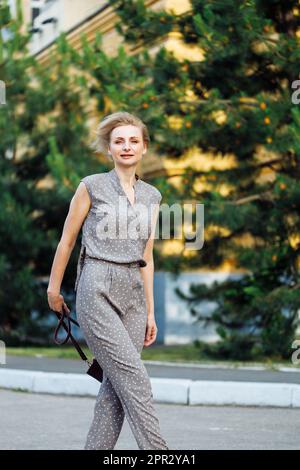 Ritratto vista laterale di una donna di 40 anni che cammina all'aperto tra abeti verdi con borsa a spalla in pelle di borgogna in mano. Signora di mezza età in bella Foto Stock