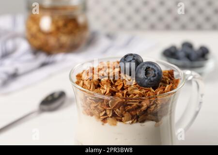 Gustoso yogurt con muesli e mirtilli in tazza servito su tavola bianca, primo piano Foto Stock