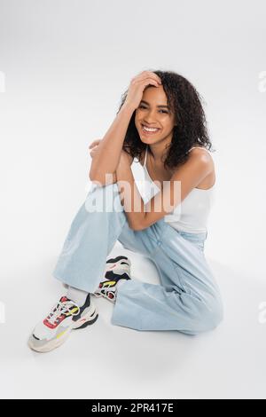 intera lunghezza di felice ragazza afro-americana in jeans blu seduta con mano vicino alla testa su sfondo grigio, immagine di scorta Foto Stock