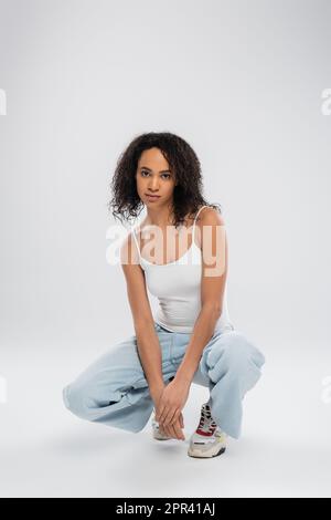 intera lunghezza di donna afroamericana in canotta bianca e jeans con sneakers seduta su hunches su sfondo grigio, immagine stock Foto Stock