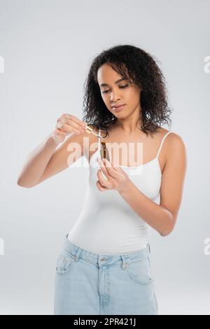 brunette afroamericana donna in possesso di contagocce e bottiglia di siero cosmetico isolato su grigio, immagine stock Foto Stock