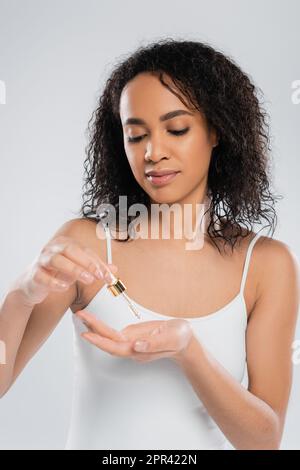 giovane afroamericana donna in bianco serbatoio top holding contagocce con siero cosmetico isolato su grigio, immagine stock Foto Stock