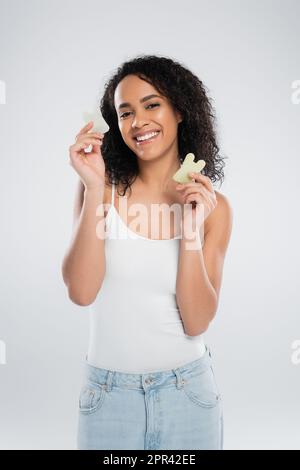 brunette afroamericana donna che tiene scraper di pietra e sorridente alla macchina fotografica isolato su grigio, immagine stock Foto Stock