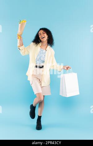 intera lunghezza di donna afro-americana overjoyed in shorts e stivali neri posando con tulipani gialli e borse della spesa su blu, immagine stock Foto Stock