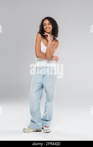 intera lunghezza di donna afroamericana gioiosa in canotta bianca e jeans blu su sfondo grigio, immagine stock Foto Stock