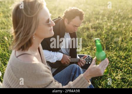 Ora legale. Bella giovane coppia godendo del tempo in campo intorno all'erba. Stile di vita, amore, datazione, concetto di vacanza. Privato. Donna che beve birra, uomo Foto Stock