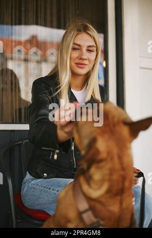 Giovane donna bionda che dona al suo cane una delizia. Allegro proprietario che alimenta il bulldog con spuntini. Foto Stock