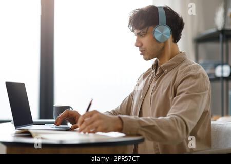 Uomo arabo che usa il laptop e che prende appunti indossando le cuffie al coperto Foto Stock