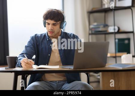 Guy prendendo appunti imparare online al laptop indossando cuffie indoor Foto Stock