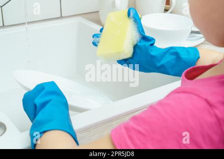 Foto ritagliata di una bambina che indossa una T-shirt rosa, guanti di gomma blu grandi, in piedi vicino al lavandino, tenendo una spugna gialla e sapone, lavando piatti dentro Foto Stock