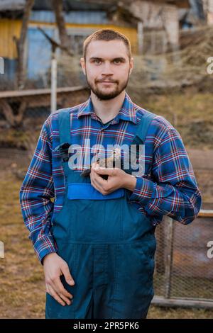 Un uomo coltivatore soddisfatto di aspetto caucasico europeo, portarato, in una camicia e tute, tiene un pollo nano in mano sullo sfondo di Foto Stock