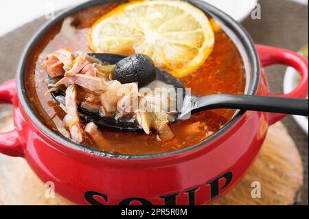 Solyanka di carne. Caldo combinato hodgepodge. una porzione di zuppa di carne rossa piccante con pomodori, salsicce affumicate e olive al limone in una turena rossa. Foto Stock
