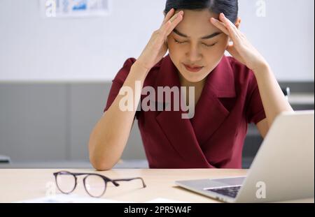 La giovane donna d'affari asiatica tiene il suo tempio con entrambe le mani, seduto in ufficio. Il concetto di disoccupazione, tristezza, problemi depressi e umani. Foto Stock