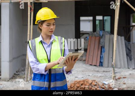 Giovane ingegnere che indossa un casco da costruzione e un giubbotto di sicurezza che controlla il programma di lavoro su un tablet computer. Ambiente di lavoro nel cantiere di progetti abitativi. Foto Stock