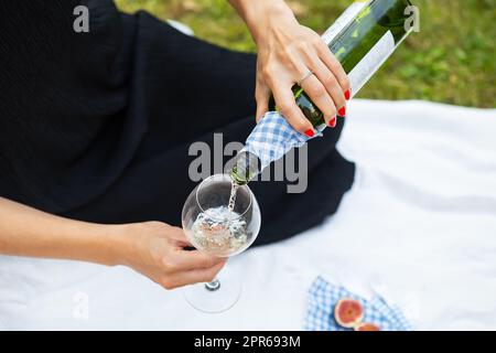 Picnic romantico nel parco sull'erba, cibo delizioso: Cestino, vino, uva, fichi, formaggio, tovaglia a scacchi blu, due bicchieri di vino. La ragazza versa vino in un bicchiere. Concetto di svago all'aperto. Foto Stock