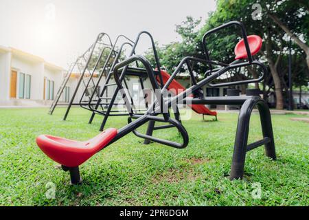 Vista mare rossa nel parco giochi. Parco giochi per bambini. Altalena in plastica o teeter-totter, altalena e scivolo nel parco giochi all'aperto con terreno erboso verde. Parco giochi all'aperto per bambini. Foto Stock