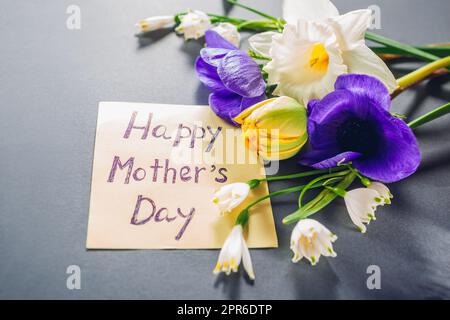 Nota di testo Happy Mother's Day con fiori primaverili. Biglietto d'auguri su sfondo grigio con fiori viola bianchi. Vista dall'alto di Flat Lay Foto Stock