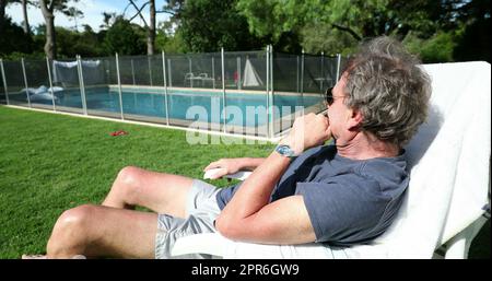 L'uomo più anziano si è seduto all'esterno riposando che guarda alla piscina nel cortile di casa. Anziano che gode di pensione Foto Stock