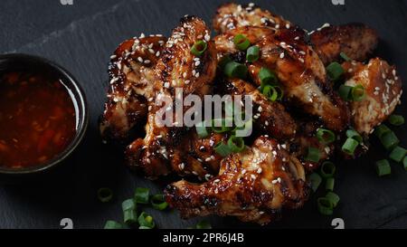 Ali di pollo alla griglia con salsa guarnita con cipolle verdi su tavola di ardesia su sfondo di pietra nera. Foto Stock