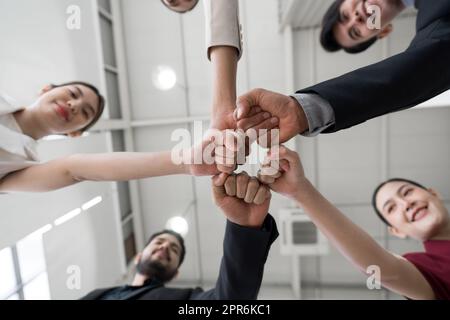 Concetto di team building. I colleghi in piedi in cerchio si scontrano a pugni, celebrando il successo aziendale in un ufficio moderno con soffitto alto. Vista inferiore Foto Stock