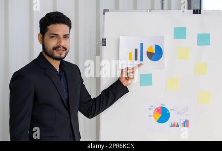 Giovane uomo d'affari con capelli ondulati, baffi e barba in abito nero puntando il dito sul grafico a torta sulla tavola bianca. Concetti di formazione e istruzione. Foto Stock