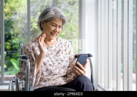 Vecchia donna asiatica con i capelli grigi che salta la mano tramite comunicazione remota tramite telefono cellulare mentre è seduta su una sedia a rotelle di fronte al corridoio con parete di vetro. Foto Stock