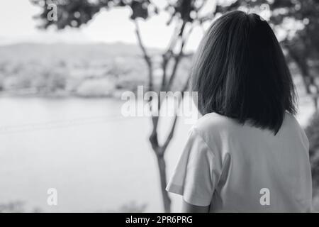 Vista sul retro della triste donna asiatica nel parco, vista sul lago. Donna depressa. Concetto di salute mentale. Donna preoccupata e ansiosa. Vita infelice. Persona sola e solitaria. Fotografia in bianco e nero. Foto Stock