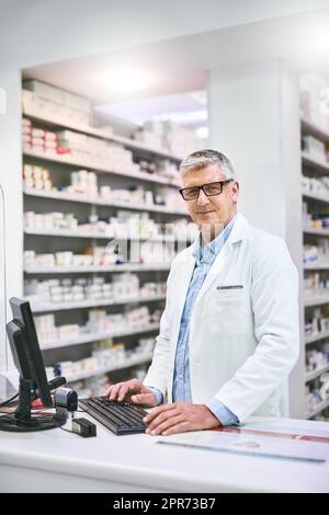 Tanti meds tra cui scegliere. Ritratto di un farmacista maschio sicuro matura digitando su un computer mentre si guarda la fotocamera in farmacia. Foto Stock