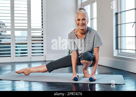 Iniziare con una semplice posa. Ritratto di una donna matura allegra che pratica yoga mentre fa uso di pesi all'interno di uno studio durante il giorno. Foto Stock