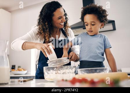 Adorabile ragazzino con afro che prepara in cucina a casa con sua madre. Allegra donna mista che setaccia la farina e mescola gli ingredienti con l'aiuto di suo figlio. La cottura è un'attività di incollaggio Foto Stock