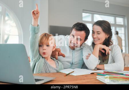 Adorabile piccola ragazza caucasica seduta al tavolo con un laptop wireless e facendo i compiti mentre la mamma e il papà la aiutano. Bella e felice giovane donna che sorride e insegna a sua figlia a casa mentre suo marito punta nella lounge di casa Foto Stock