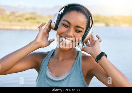 Donna eccitata che sorride e ascolta musica con l'acqua in sottofondo. Felice giovane donna che ascolta i brani della sua playlist in streaming online mentre si trova all'aperto. Utilizzando le cuffie wireless, è possibile riprodurre brani musicali Foto Stock