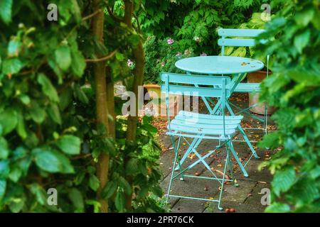 Sedie da giardino e tavolo in un cortile privato, tranquillo, lussureggiante e sereno. Set di mobili da patio in metallo e posti a sedere in un cortile vuoto e tranquillo. Luogo tranquillo e rilassante con fiori e piante fresche Foto Stock