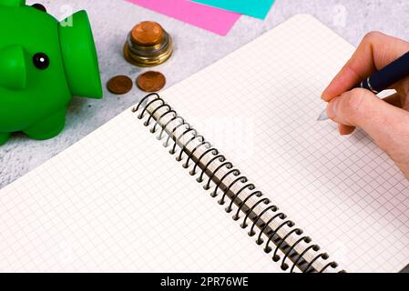 Uomo che scrive un messaggio importante sul notebook sulla scrivania con denaro, penna, appunti e maiale. Informazioni critiche sul blocco note sul tavolo con centesimi, Memo e matita. Foto Stock