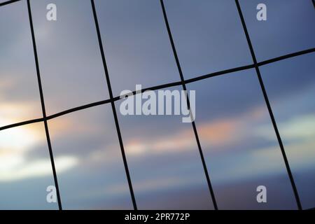 Stirare la recinzione in linee verticali orizzontali sullo sfondo del cielo sfocato. Filo intrecciato in forma rettangolare per un concetto di sicurezza o sicurezza. La texture dei motivi delle finestre o delle silhouette scure di un cancello Foto Stock